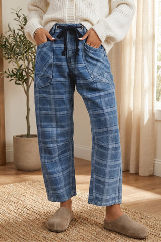 A person wearing blue plaid cropped pants with a drawstring waist, paired with a cream knit sweater and taupe slip-on shoes, standing indoors on a woven rug near a window with curtains.