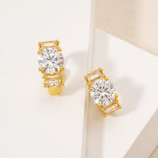 A pair of gold-toned stud earrings with a large round clear center stone and three baguette-cut clear side stones, displayed on a clean white geometric surface.