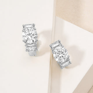 A pair of silver-toned stud earrings with baguette and round-cut clear gemstones, displayed on a clean cream-colored geometric surface.