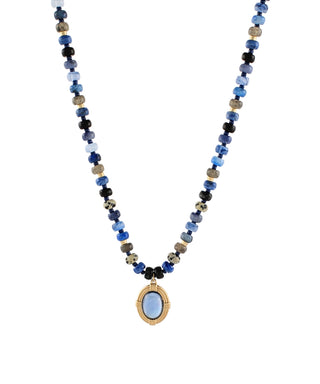 A beaded necklace featuring a mix of blue, black, and metallic gold beads, with a central oval pendant containing a light blue stone surrounded by a textured gold border, set against a plain white background.