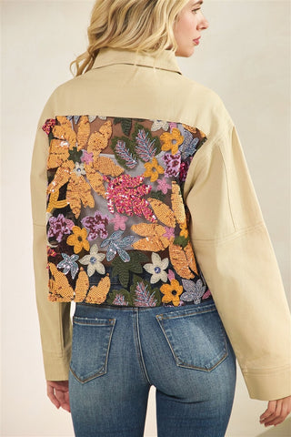Person wearing a beige jacket with a colorful floral and butterfly pattern on the back, standing against a neutral background.