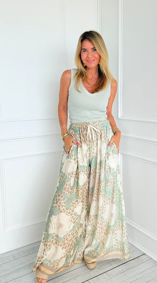 A woman modeling a sleeveless light sage green top and matching wide-leg maxi pants with a beige and green floral print, standing in a bright white room with white paneled walls and light wood flooring.
